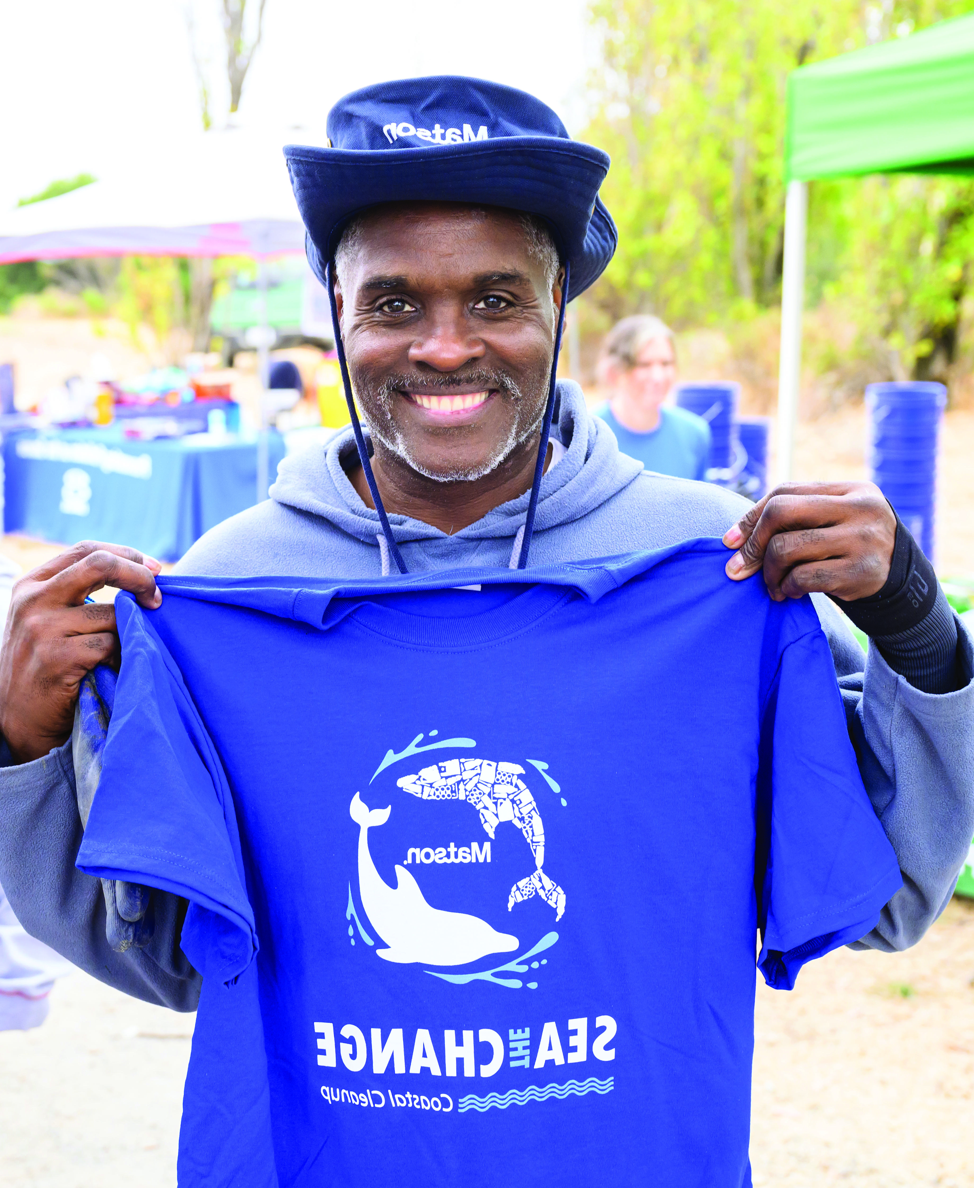 Volunteer wears a blue 澳博体育app下载 branded hand and holds up a blue t-shirt printed with two dolphins encircling a 澳博体育app下载 logo and the words "Sea The Change."