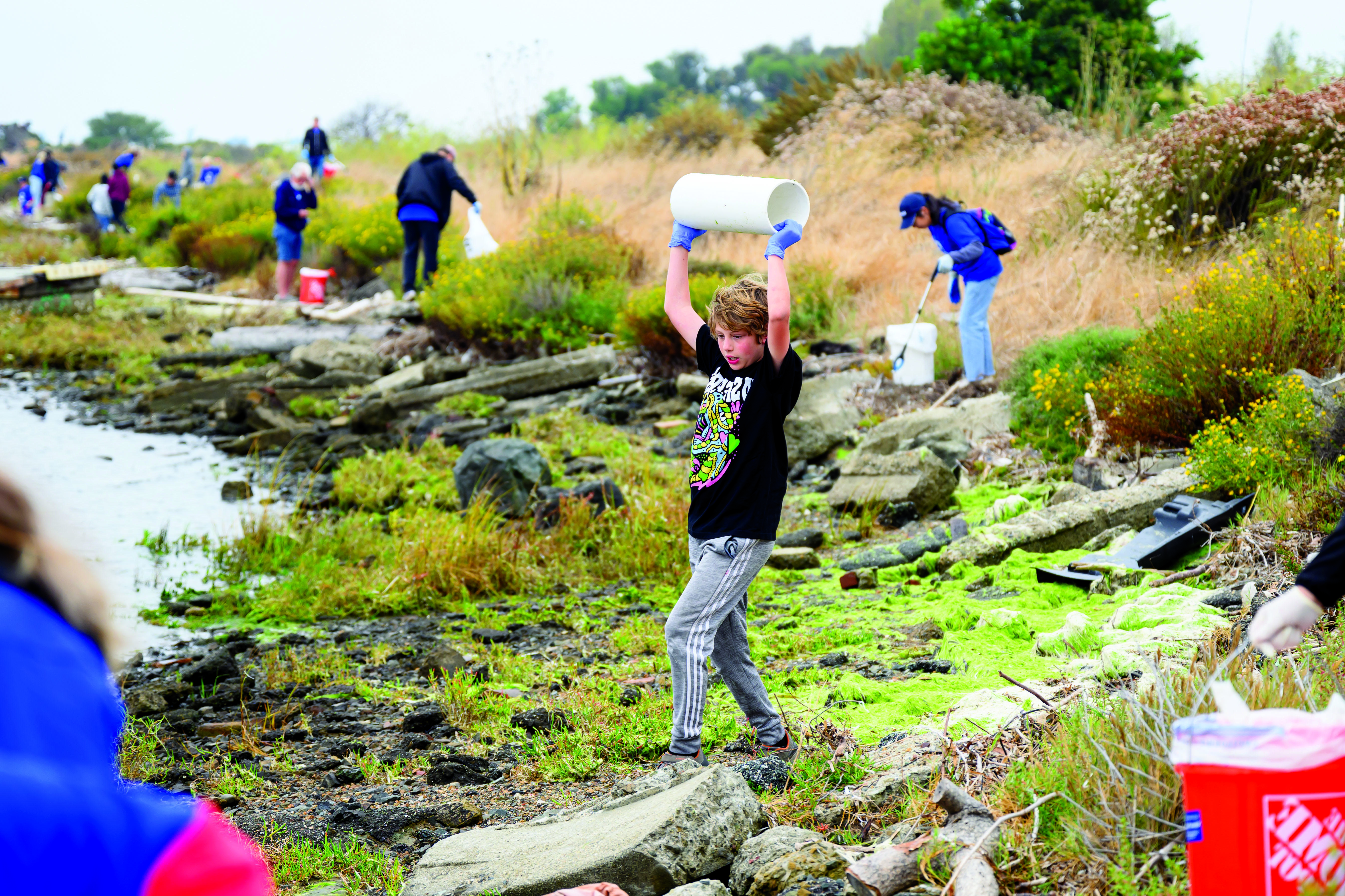 Young male tween wearing blue plastic gloves holds up a large section of plastic piping at the estuary wtih other clean up volunteers in the background.